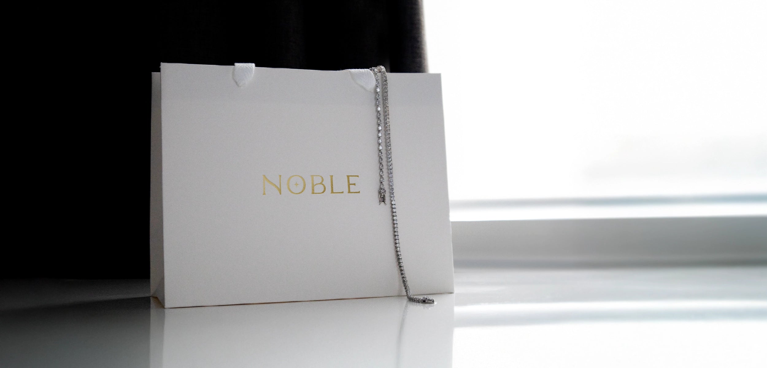 A white Noble gift bag with gold foil logo sitting on a white surface with a tennis necklace and bracelet draped over